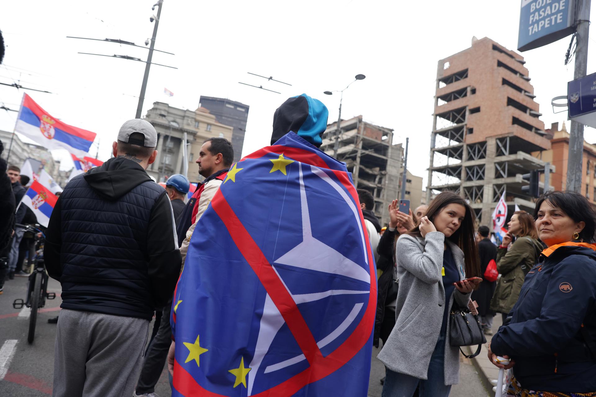 Un manifestante, envuelto en una bandera antimperialista de la OTAN y de la UE, asiste a la protesta en el sitio del antiguo edificio del Estado Mayor para conmemorar el 26.º aniversario de la campaña de bombardeo de la OTAN de 1999 en Belgrado, Serbia, 24 de marzo de 2025. La manifestación se produce en medio de la controversia sobre un acuerdo que otorga a la firma de inversiones de Jared Kushner un contrato de arrendamiento de 99 años para desarrollar un complejo de lujo y un museo en el lugar. (Protestas, Belgrado) EFE/EPA/ANDREJ CUKIC