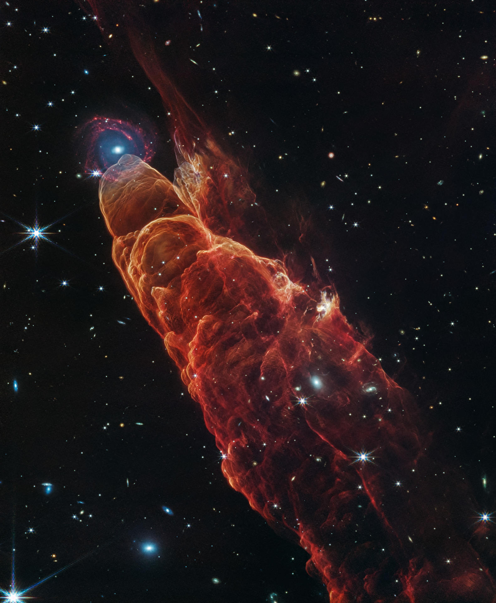 Imagen tomada con los instrumentos NIRCam y MIRI del telescopio espacial James Webb. La nube de forma cónica de color rojo anaranjado corresponde a Herbig-Haro 40/50, en el extremo superior izquierdo hay una galaxia de fondo en forma de espiral con un centro azul. EFE/Crédito: NASA, ESA, CSA, STScI/SOLO USO EDITORIAL