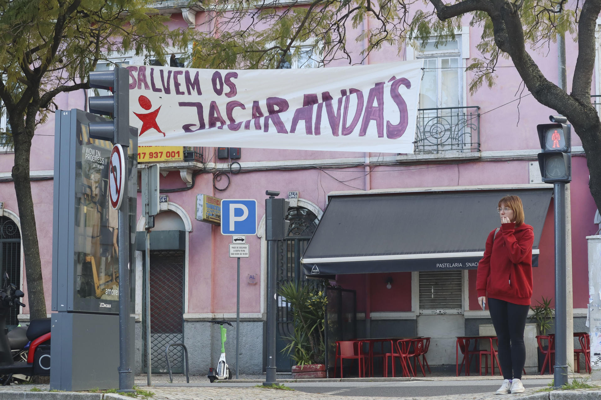 Una joven pasea este martes bajo un cartel que denuncia la tala y el trasplante de jacarandás previstos en la Avenida 5 de Outubro, en el centro de Lisboa, para construir un aparcamiento subterráneo. EFE/Carlos Caselles