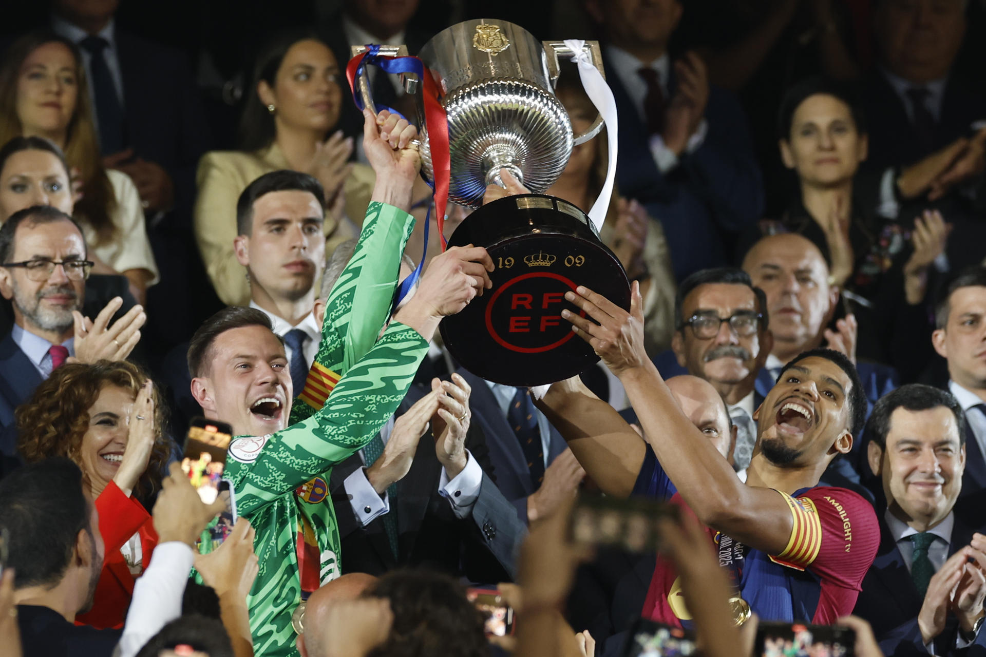 Los capitanes del Barcelona Marc Andre Ter Stegen (i) y Ronald Araujo sostienen el trofeo de la Copa del Rey tras la final que disputaron Real Madrid y FC Barcelona en el estadio de La Cartuja, en Sevilla. EFE/Julio Muñoz
