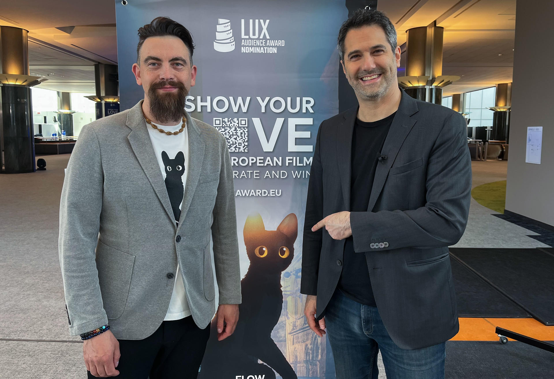 El coproductor del filme Flow, Grégory Zalcman (d), y el compositor de la Banda sonora, Rihards Zalupe, posan frente al póster de la película en una entrevista con EFE momentos previos a la gala de los Premios Lux del Parlamento Europeo, celebrada este martes en Bruselas. EFE/ Sara Morato