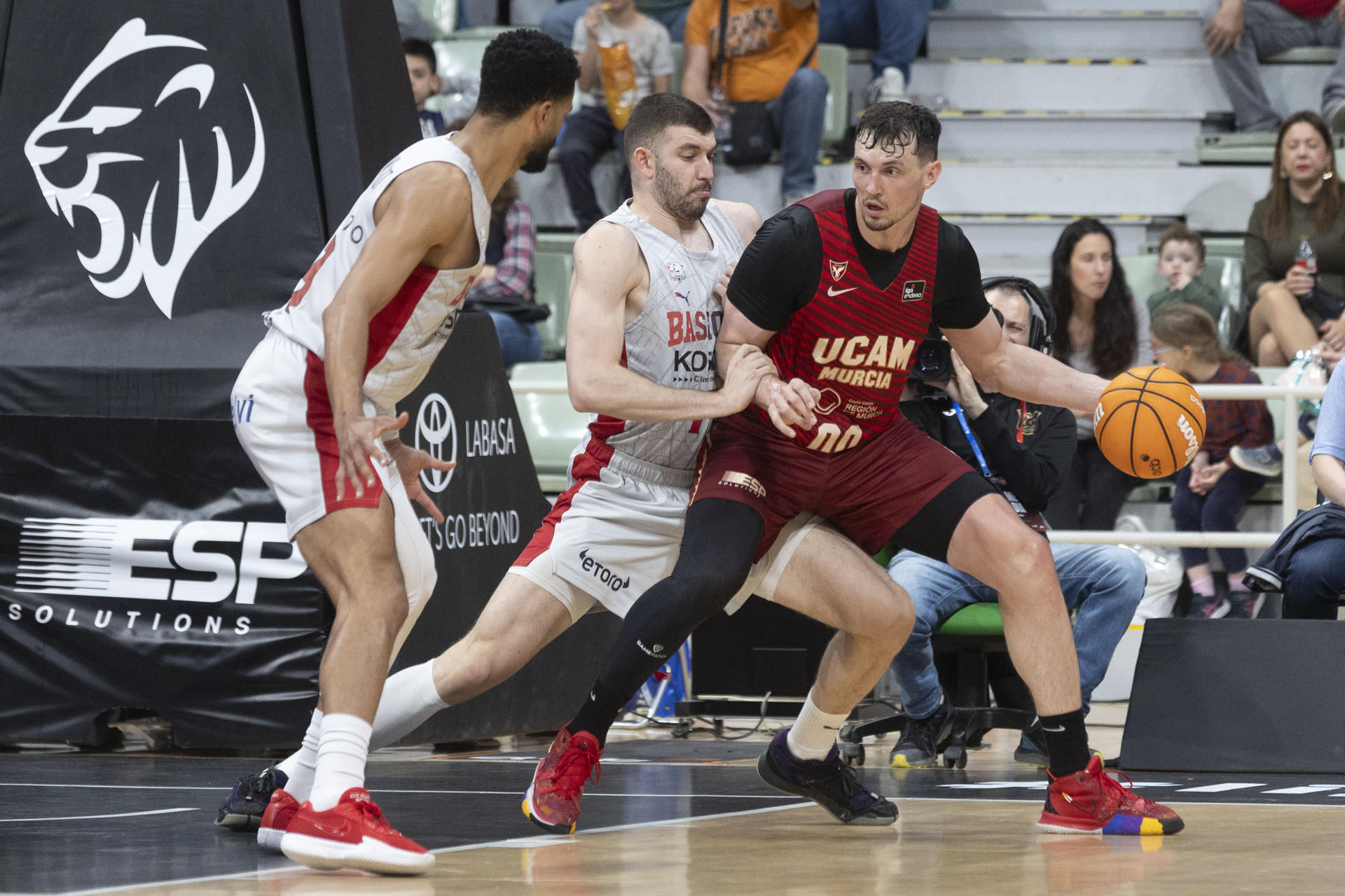 El alero letón de UCAM Murcia Aturcs Kurucs (d) juega un balón ante la presión del alero griego del Baskonia Nikos Rogkavopoulos (c), durante el partido de la vigésimo séptima jornada de la liga Endesa jugado en el Palacio de los Deportes de Murcia. EFE/Marcial Guillén