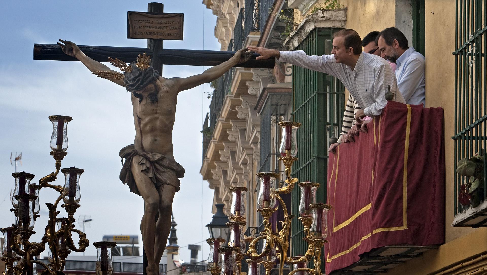 Imagen de archivo de un hombre asomado a un balcón alcanza a tocar la mano del Cristo de la Salud de la Hermandad de San Bernardo a su paso por calles céntricas de la ciudad de Sevilla. EFE/José Manuel Vidal