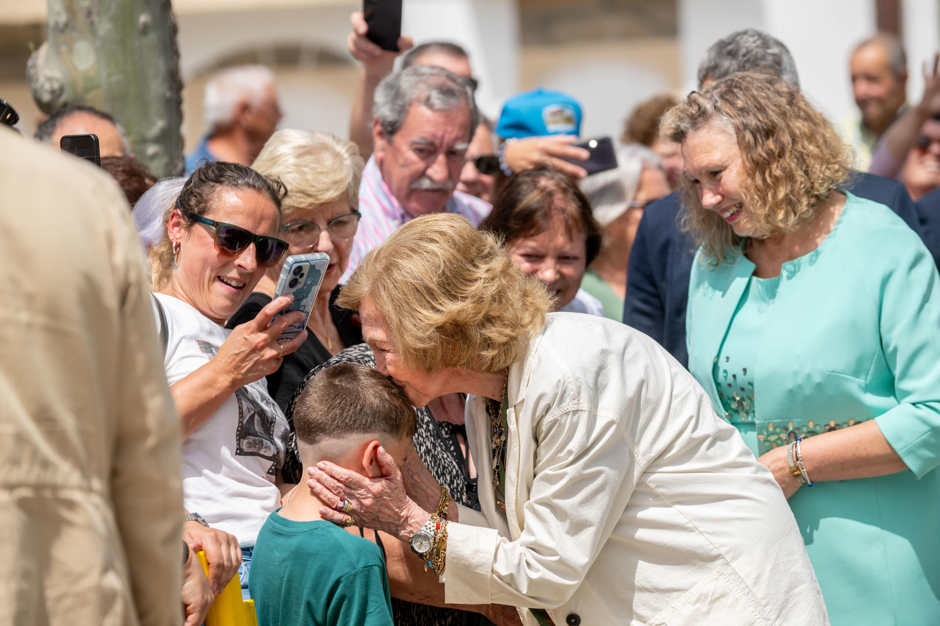 La reina emérita Sofía, acompañada por la alcaldesa de la localidad, Sara Inés Vega (d), saluda a un niño mientras visita Castro Caldelas (Ourense) este viernes como parte de una iniciativa de su Fundación orientada a promover el valor sostenible del medio rural y su patrimonio cultural y natural. EFE/ Brais Lorenzo