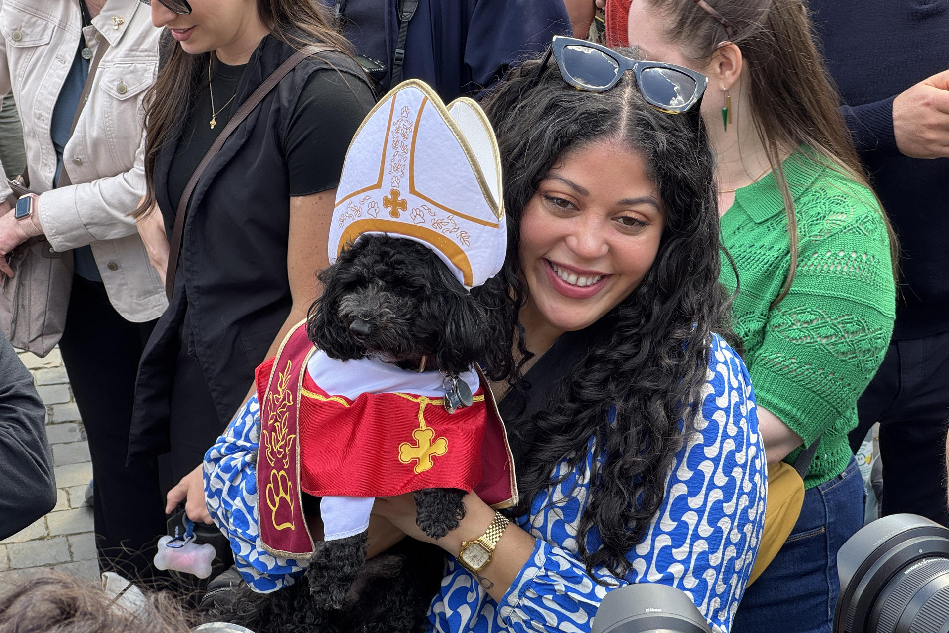 Una mujer junto a su perro disfrazado de Papa es fotografiada por la prensa, en la plaza de San Pedro, este jueves. Unas 11.000 personas se congregaron este jueves en la plaza de San Pedro del Vaticano y su entorno para ver la segunda fumata del cónclave, que salió negra, por lo que sigue sin haberse elegido un nuevo papa, y se espera que sean muchas más las que se den cita de nuevo en el lugar para la fumata de la tarde. EFE/ Daniel Cáceres