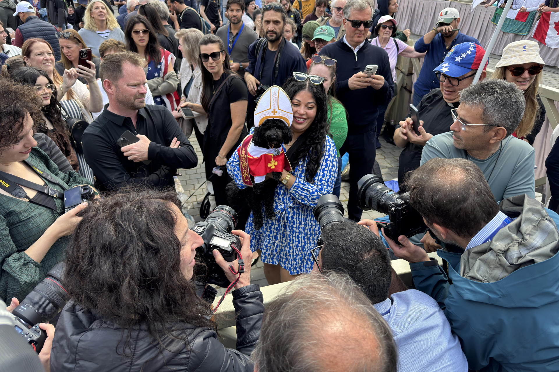 Una mujer junto a su perro disfrazado de Papa es fotografiada por la prensa. Unas 11.000 personas se congregaron este jueves en la plaza de San Pedro del Vaticano y su entorno para ver la segunda fumata del cónclave, que salió negra, por lo que sigue sin haberse elegido un nuevo papa, y se espera que sean muchas más las que se den cita de nuevo en el lugar para la fumata de la tarde. EFE/ Daniel Cáceres