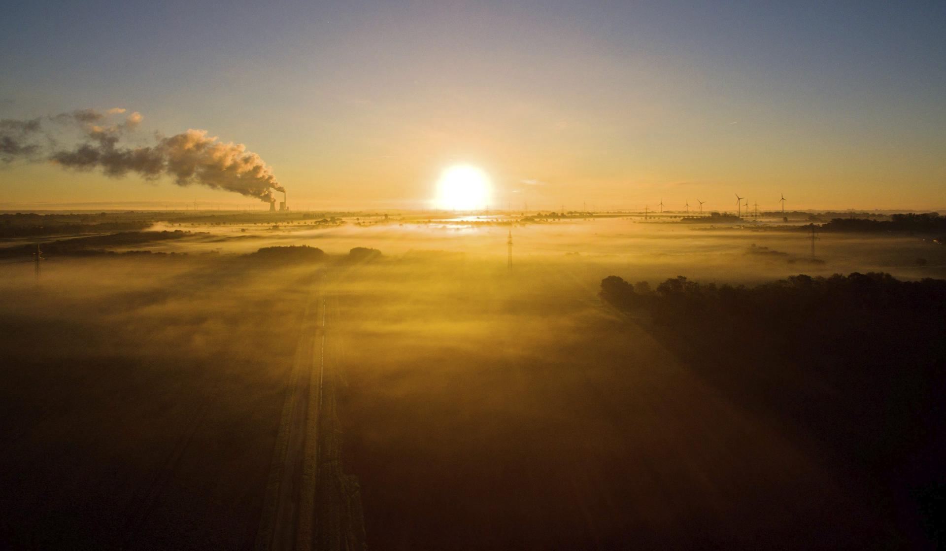 El sol se asoma en el horizonte al amanecer en un campo eólico con el humo de una fábrica de fondo, en una imagen de archivo. EFE/Julian Stratenschulte