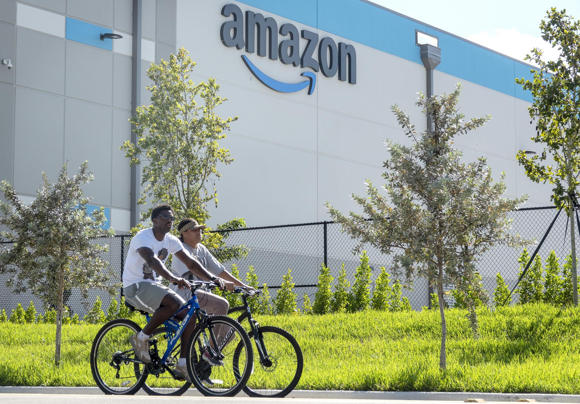 Fotografía de archivo de dos personas circulando en bicicleta frente a un centro logístico de Amazon en Florida (EE.UU.). Zoox, la empresa de vehículos autónomos propiedad de Amazon, inauguró una planta que se centrará en la producción de robotaxis en California, con la que busca hacer competencia a Waymo, según apuntan este miércoles los medios locales. EFE/EPA/ CRISTOBAL HERRERA-ULASHKEVICH / ARCHIVO