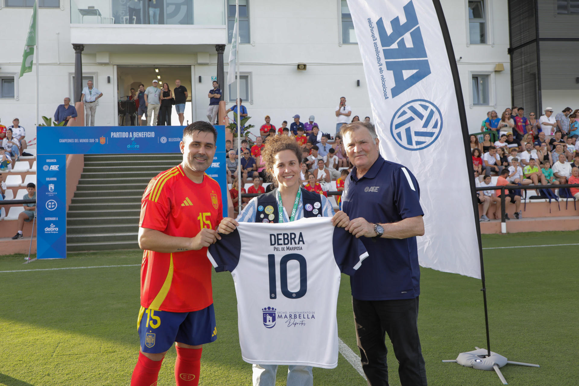 El presidente de AFE, David Aganzo, (izq), y el exfutbolista y entrenador José Antonio Camacho entregan una camiseta una camiseta conmemorativa a la coordinadora de DEBRA Piel de Mariposa, Natividad Romero, como muestra de apoyo a la labor social que desarrolla la organización en favor de las personas afectadas por esta enfermedad rara, durante el partido benéfico disputado en Marbella este martes. EFE/AFE