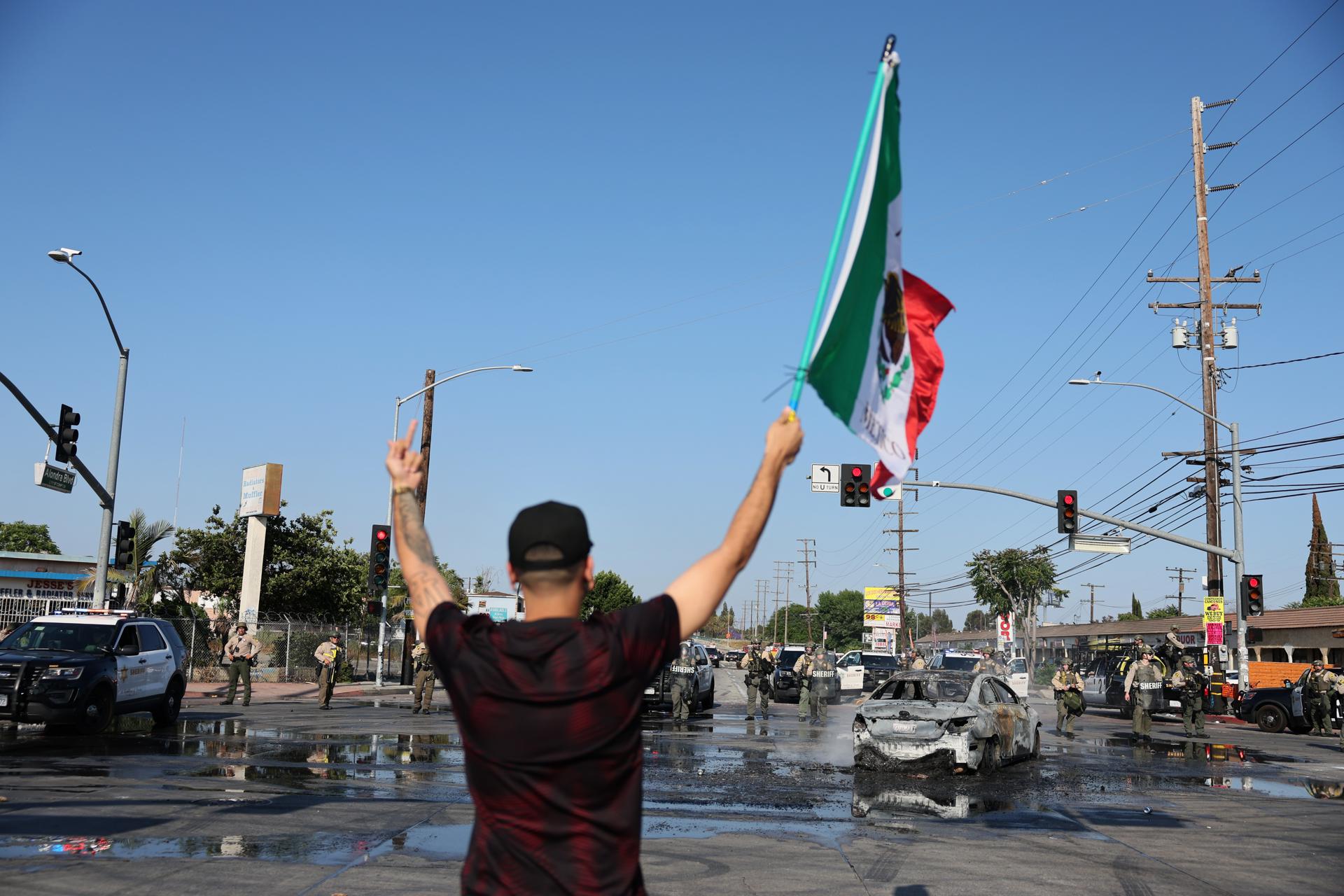 Un manifestante fue captado este sábado, 7 de junio, al sostener una bandera mexicana cerca de un automóvil quemado, durante un enfrentamiento entre manifestantes y agentes federales cerca después de una redada realizada por el Servicio de Inmigración y Control de Aduanas (ICE) en contra presuntos migrantes indocumentados, en Paramount (California, EE.UU.). EFE/Allison Dinner