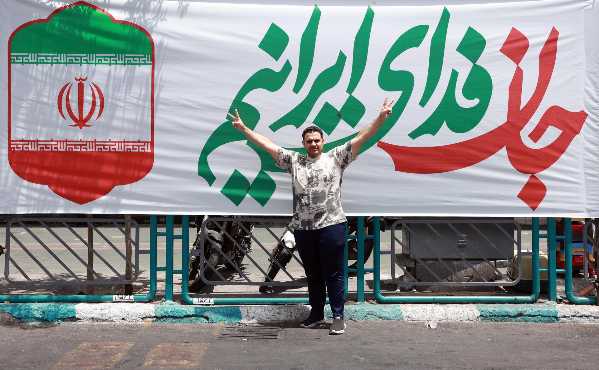 Un iraní hace la señal de la victoria ante un cartel con la bandera de Irán junto al lema: 'Mi vida es un sacrificio por Irán', instalado en la plaza Enghelab, en el centro de Teherán, el 24 de junio de 2025. EFE/EPA/ABEDIN TAHERKENAREH