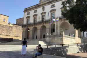 Varias personas junto al Ayuntamiento de Cáceres donde ondean las banderas del Orgullo LGTBI, este domingo. La actuación de una mujer que "por su cuenta y riesgo" ha arriado las banderas del Orgullo LGTBI izadas en el Ayuntamiento de Cáceres este domingo durante el desfile del Corpus Christi, ha llevado al propio alcalde del PP, Rafael Mateos, a denunciar la situación a la Policía Local, que ha identificado a la mujer in situ. EFE/ Eduardo Villanueva