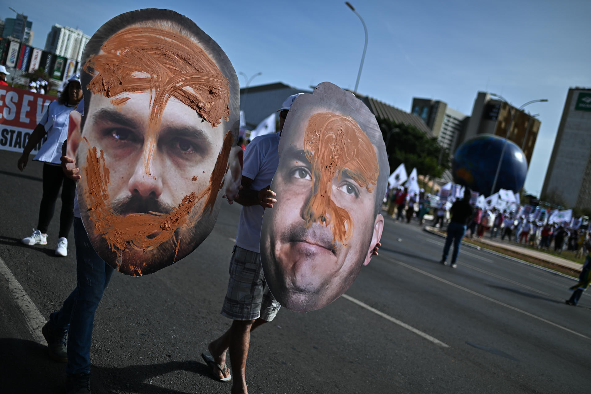 Personas sostienes carteles manchados de barro con imágenes del diputado en licencia Eduardo Bolsonaro (i) y del senador Flávio Bolsonaro (d), hijos del expresidente Jair Bolsonaro, durante una manifestación por el Día del Medio Ambiente y contra el proyecto de ley "de la devastación" que propone cambios al sistema de licencias ambientales en la Explanada de los Ministerios, este jueves, en Brasilia (Brasil). EFE/Andre Borges
