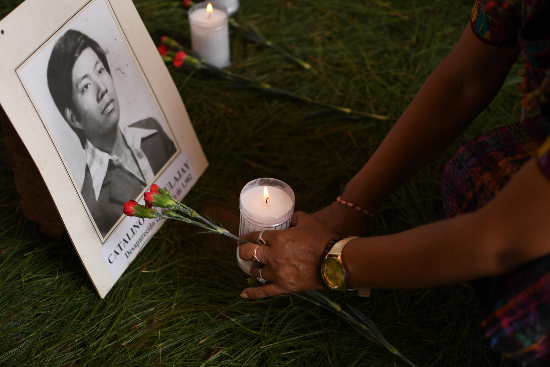 Detalle de los brazos de una mujer al colocar una vela y una flor frente al retrato de un familiar que fue víctima de desaparición forzada en Guatemala, durante un acto que honra su memoria en Ciudad de Guatemala. EFE/Alex Cruz