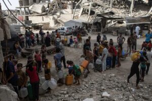 Decenas de ciudadanos palestinos desplazados fueron captados este domingo, 24 de agosto, al esperar para llenar contenedores con agua potable, entre los escombros en el campamento de Khan Younis, en la Franja de Gaza. EFE/Haitham Imad