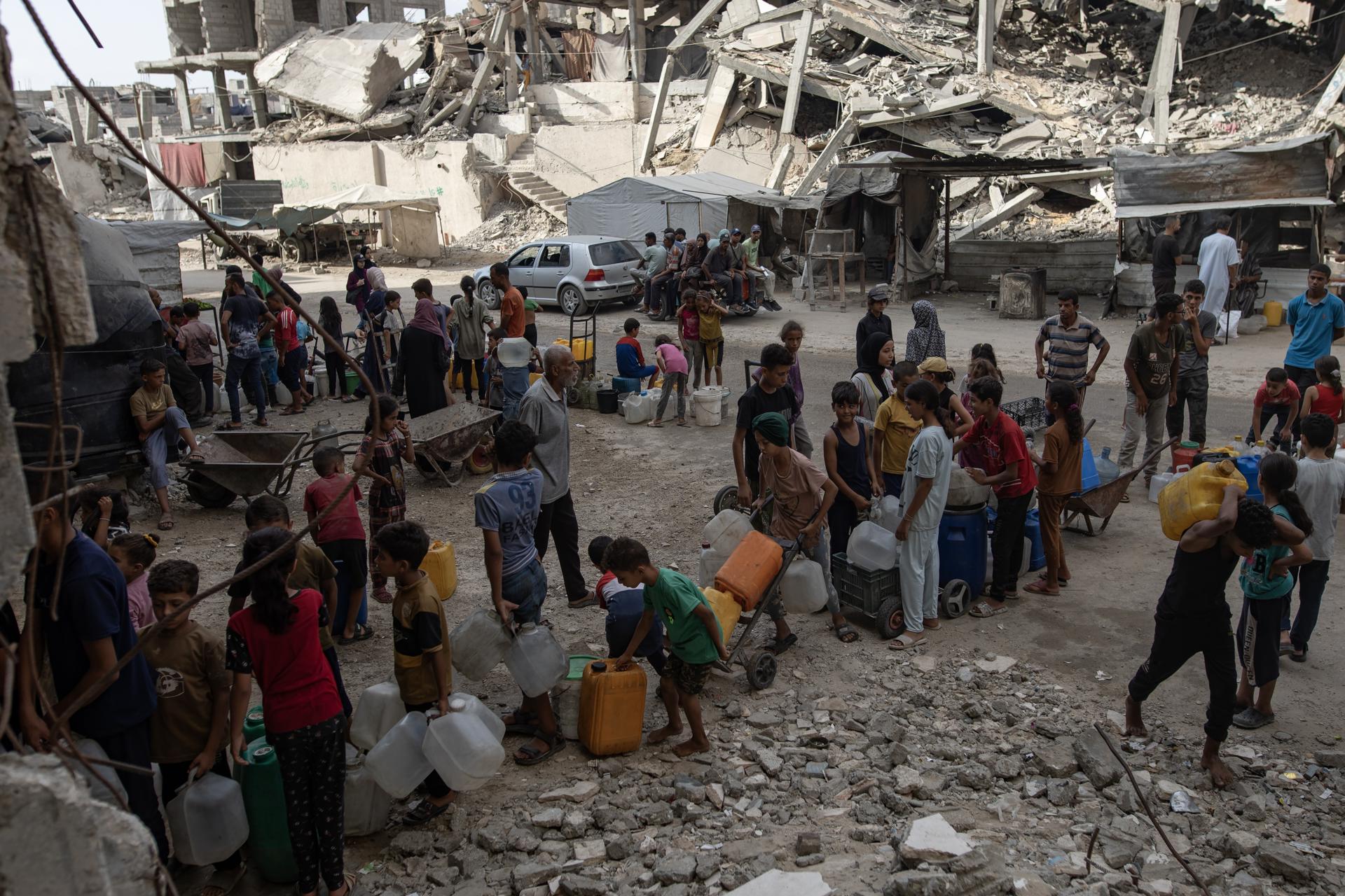 Decenas de ciudadanos palestinos desplazados fueron captados este domingo, 24 de agosto, al esperar para llenar contenedores con agua potable, entre los escombros en el campamento de Khan Younis, en la Franja de Gaza. EFE/Haitham Imad