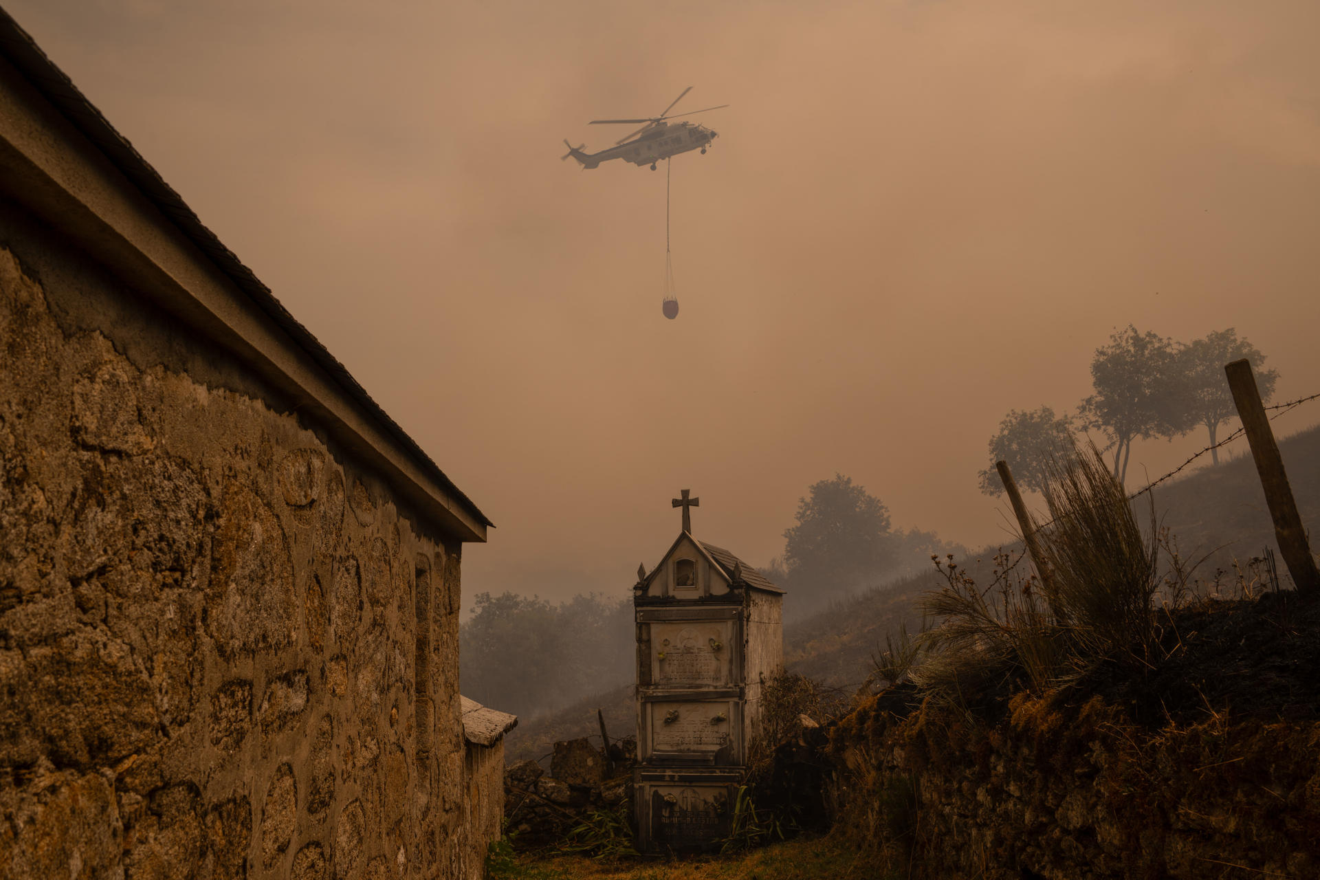 Un helicóptero de control de incendios sobrevuela el cementerio la localidad de Vilar durante el incendio forestal que permanece activo en Chandrexa de Queixa (Ourense), este martes. EFE/Brais Lorenzo
