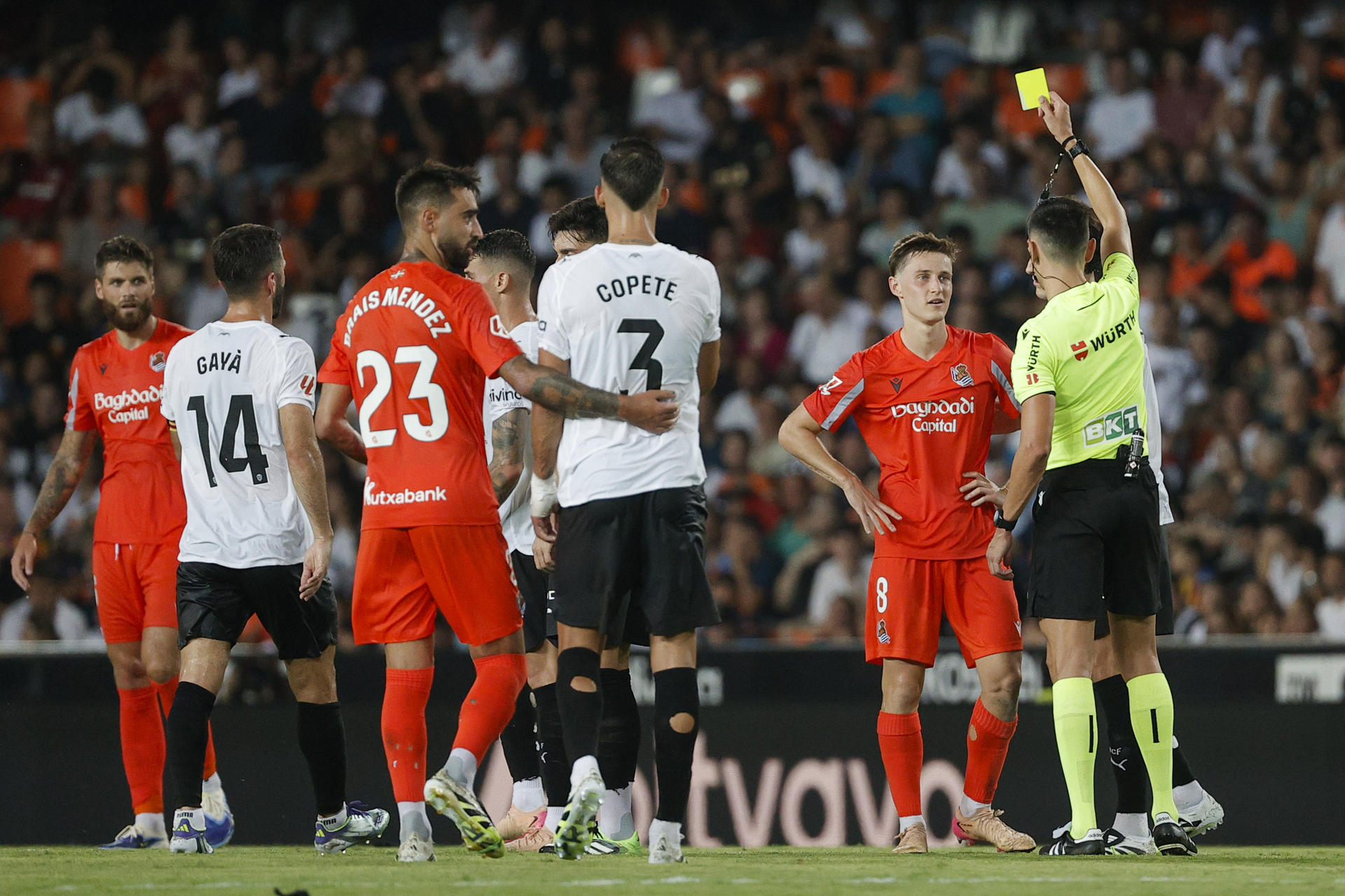 El árbitro Sánchez Martínez saca tarjeta amarilla a Beñat Turrientes (2d), de la Real Sociedad, durante el partido de la primera jornada de LaLiga entre Valencia CF y Real Sociedad, este lunes en el estadio de Mestalla, en Valencia. EFE/ Manuel Bruque
