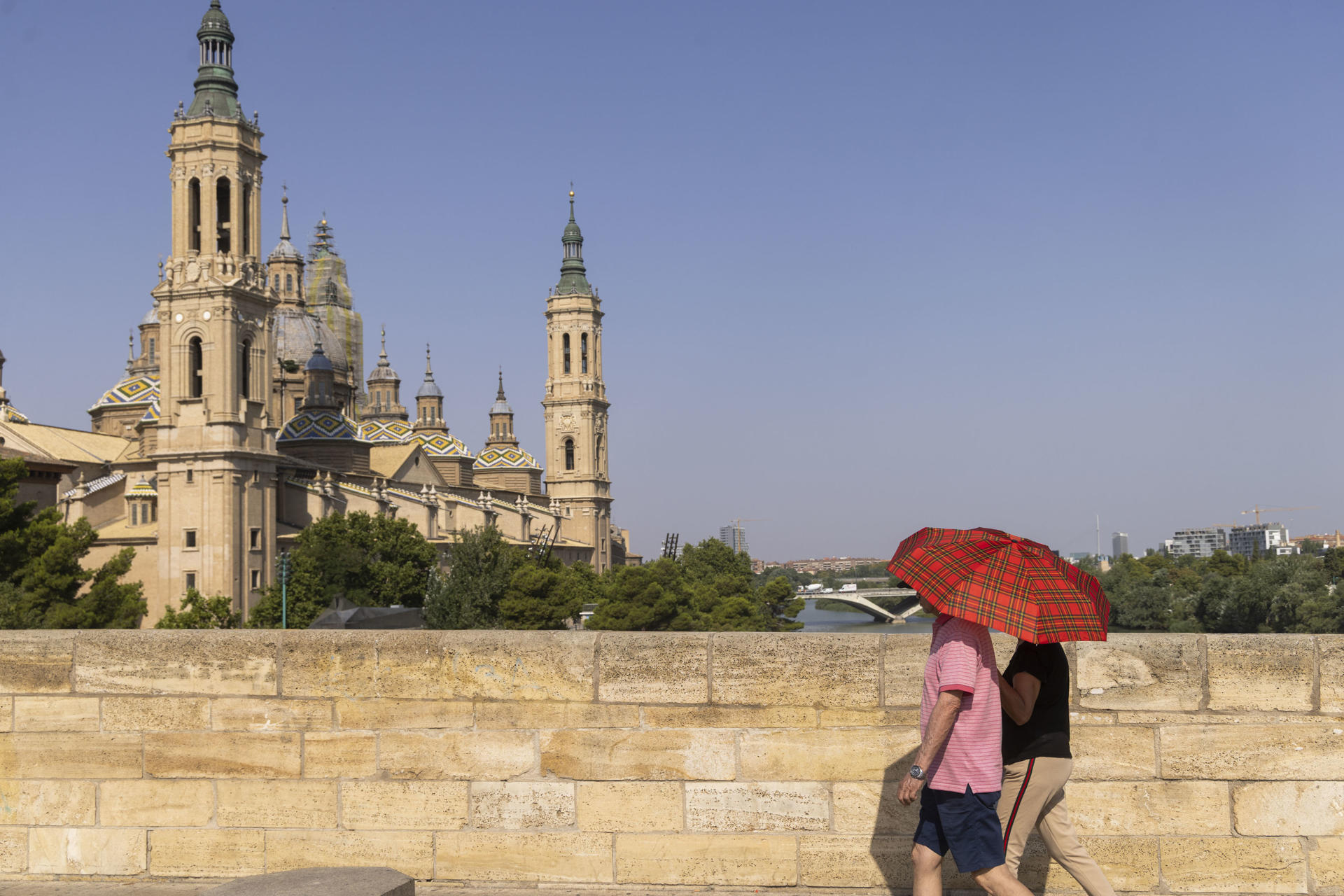 Una pareja cubierta con un paraguas para protegerse del sol pasa este jueves, por el puente de Piedra en Zaragoza. EFE/ Toni Galán