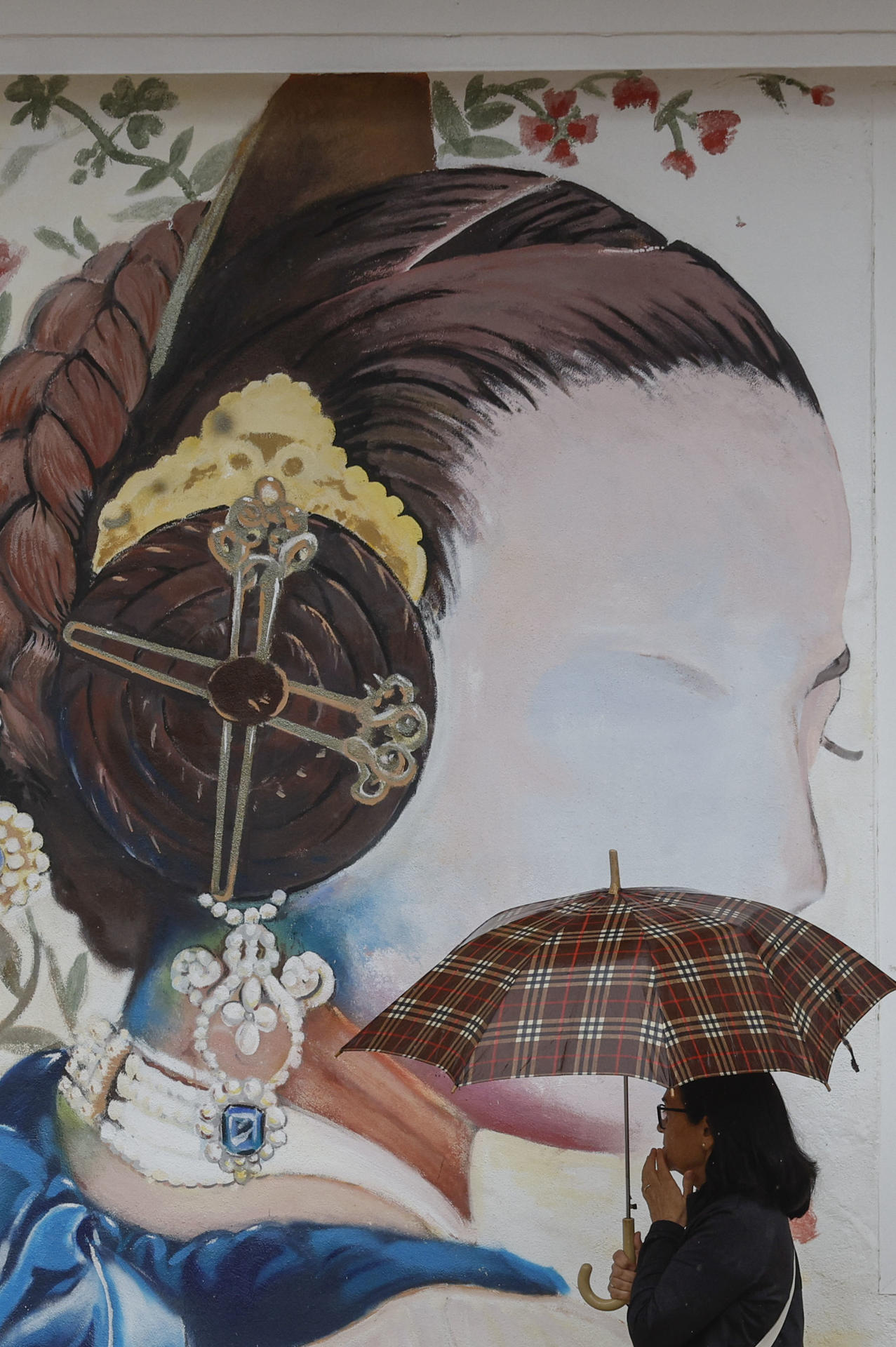 Una mujer protegida de la lluvia con un paraguas pasa ante un mural que representa a una fallera este jueves en Catarroja (València). EFE/ Kai Forsterling